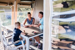 Une famille de trois personnes est assise autour d’une table en bois, un téléphone intelligent posé dessus, à l’intérieur d’une structure ressemblant à une tente, d’où l’on aperçoit un paysage verdoyant.