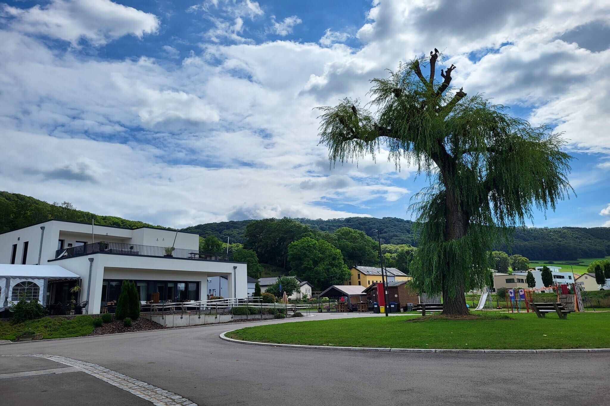 Een modern gebouw met een gebogen oprit, een grote boom ervoor en een speeltuin op de achtergrond.