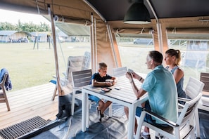 Un espace extérieur abrité avec une table et des chaises, une personne utilisant un smartphone, et en arrière-plan, une vue sur des tentes et de l’herbe.