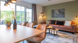 A modern living room with a sofa, coffee table, and dining table. There is a large window with curtains, a vase with flowers, and a painting on the wall. - Holiday Home near Biesbosch & Breda (Oosterhout)