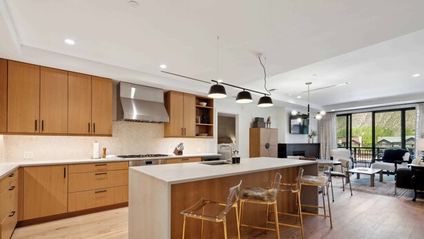 A modern kitchen with wooden cabinets, a central island with bar stools, and a dining area with a large window.