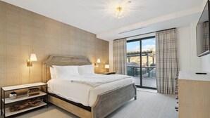 A modern hotel room with a large bed, two bedside lamps, a nightstand with books, a wooden headboard, a large window with curtains, and a TV on a wooden stand.
