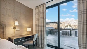 A hotel room with a bed, a desk with a chair, a lamp, and a large window with curtains.