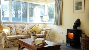A cozy living room with a sofa, a table with books, a lamp, and a wood-burning stove.