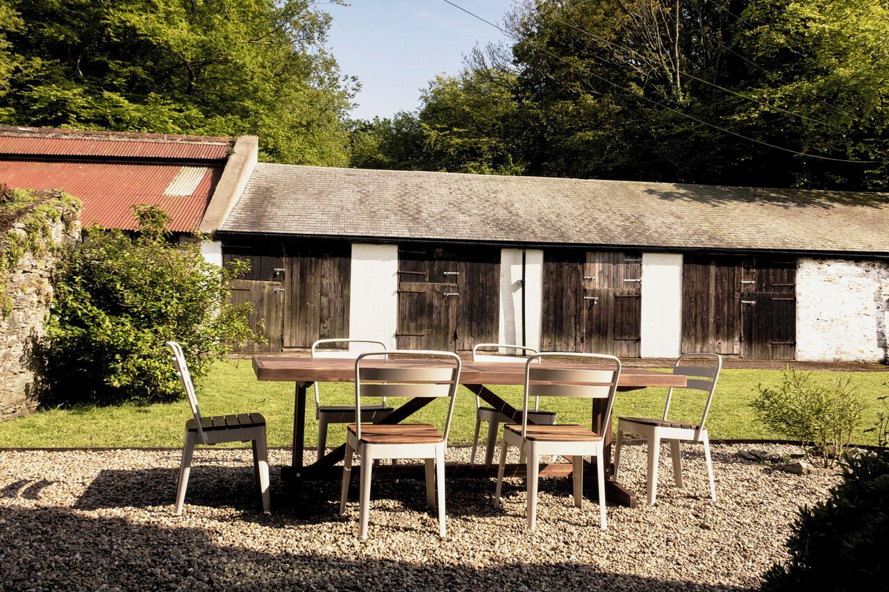 Un espace de restauration extérieur rustique, avec une table en bois et des chaises en métal, le tout mis en valeur par une grange et de la verdure en arrière-plan.