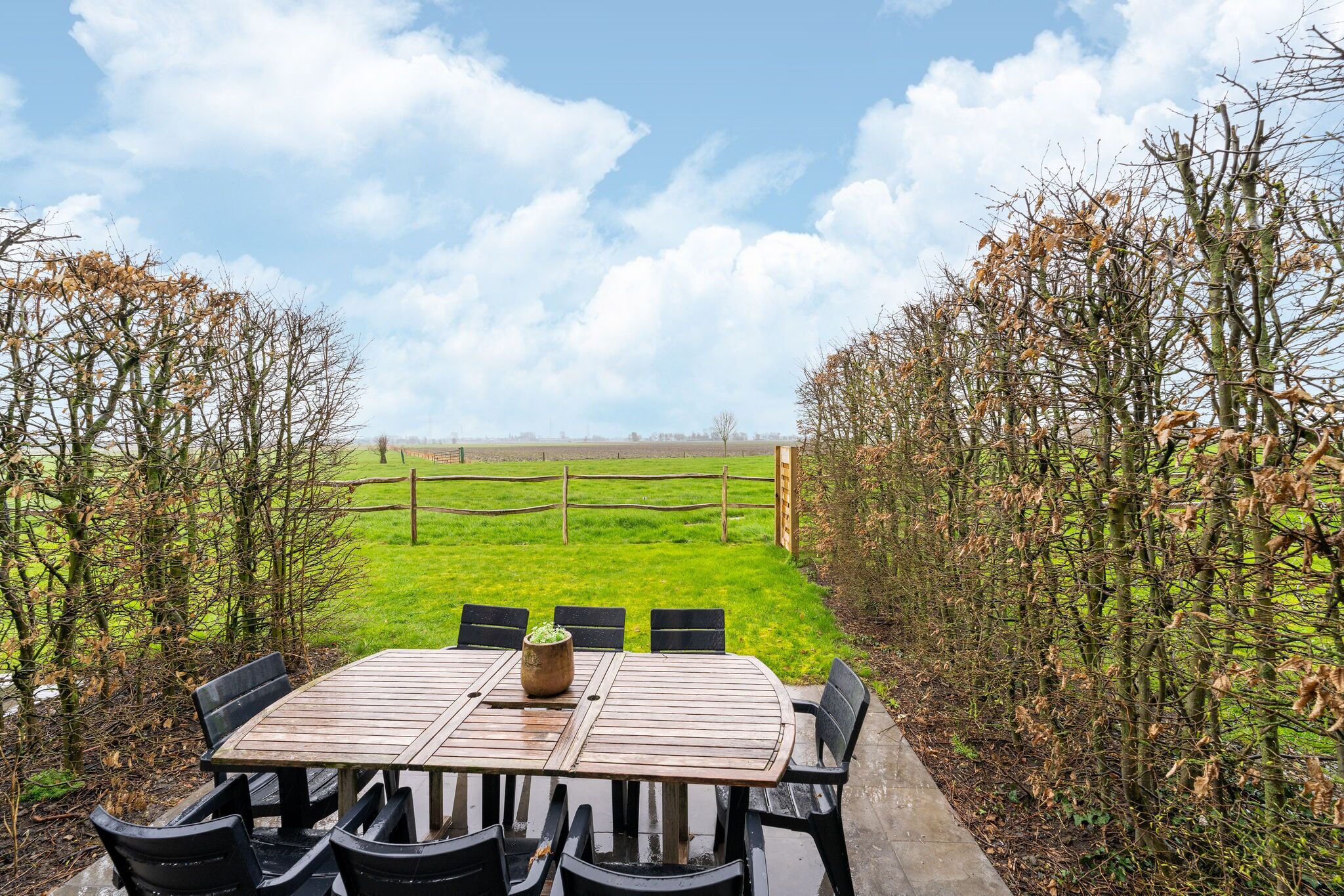 Une table en bois, entourée de chaises, est placée à l’extérieur, offrant une vue sur un champ verdoyant et un ciel dégagé.