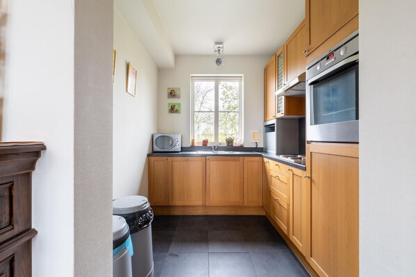 A compact kitchen with wooden cabinets, a microwave, a stove, and a window providing natural light. - Comfortable group home with large garden (Diksmuide)
