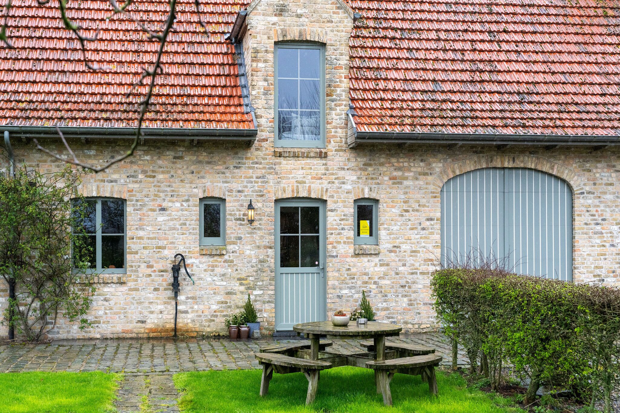 A stone building with a tiled roof, a central door, and a small garden with a wooden table and chairs.