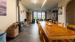 A spacious dining area with a large wooden table and chairs, a fireplace, and a view of the outdoors through large windows. - Apartment on Belgium's Stunning Coastline (Diksmuide)