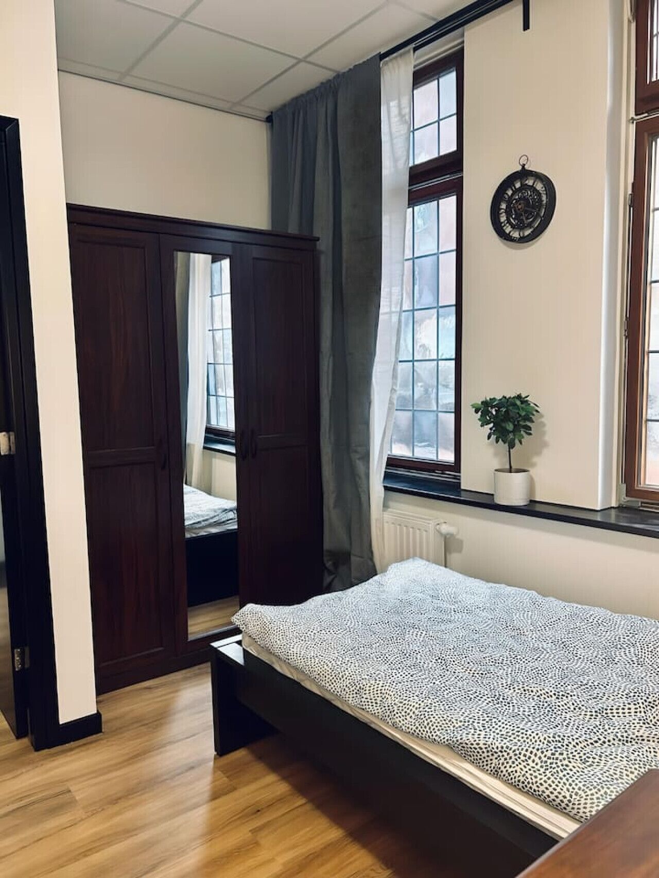 Ein Schlafzimmer mit einem Bett, einem Fenster mit Vorhängen, einem Heizkörper und einer Uhr an der Wand.
