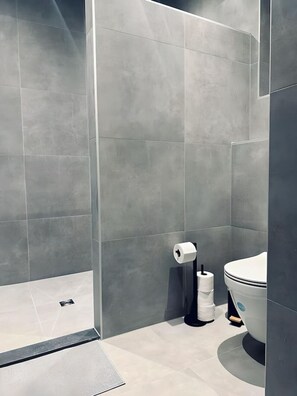 A modern bathroom with grey tiles, a toilet, a toilet paper holder, and a shower area. - Fewo Kupferkrug - Absolut Zentral + Historie (Nordhorn)
