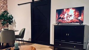 Un salon moderne avec un téléviseur diffusant une image de feu de cheminée, un meuble de rangement noir pour appareils électroniques, une table à manger avec des chaises et une porte coulissante.