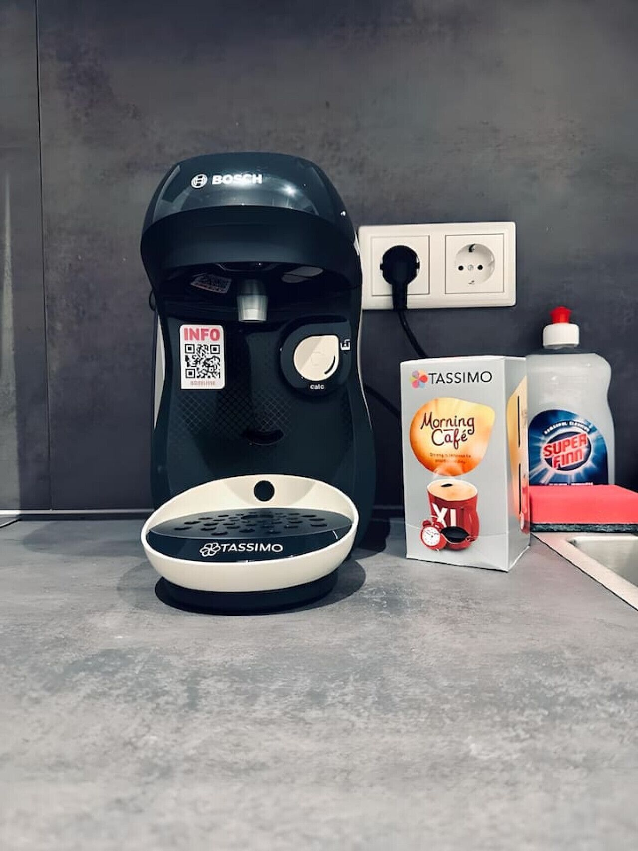 Une machine à café, une boîte de dosettes Tassimo et une bouteille de café Tassimo sont posés sur un plan de travail.