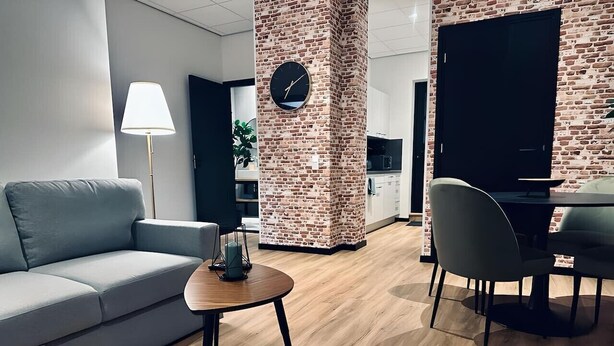 Un salon moderne avec un canapé gris, une table basse ronde en bois, un mur décoratif en briques agrémenté d’une horloge, et un coin repas avec une table et des chaises.