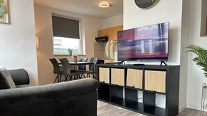 Un saló modern amb un sofà, una zona de menjador amb cadires i una taula, una cuina amb armaris i un televisor de pantalla plana gran muntat a la paret.