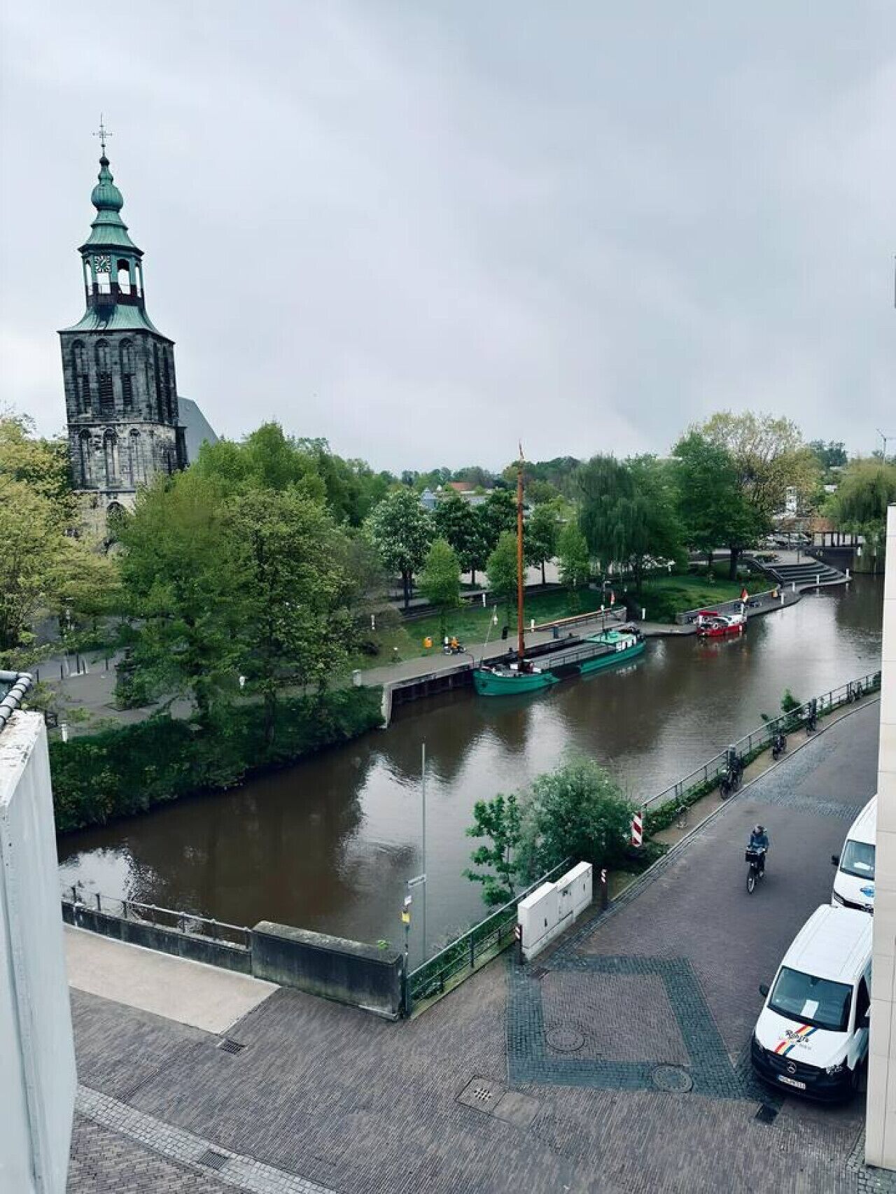A riverside view with a historic tower, a green boat docked, and a paved walkway.
