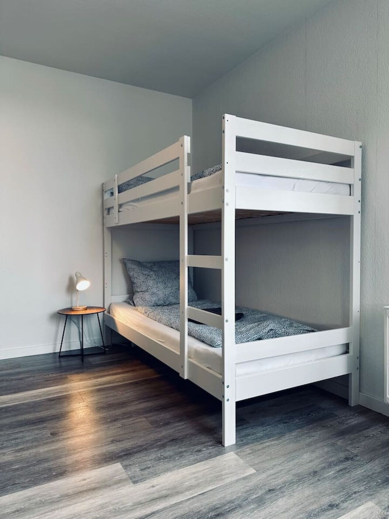 Un lit superposé avec une table de chevet et une lampe, dans une pièce au plancher en bois et aux murs de couleur gris clair.