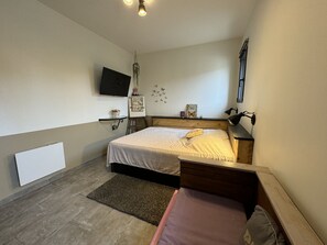 A bedroom with a bed, bedside table, lamp, wall-mounted TV, and a small shelf with decorative items.