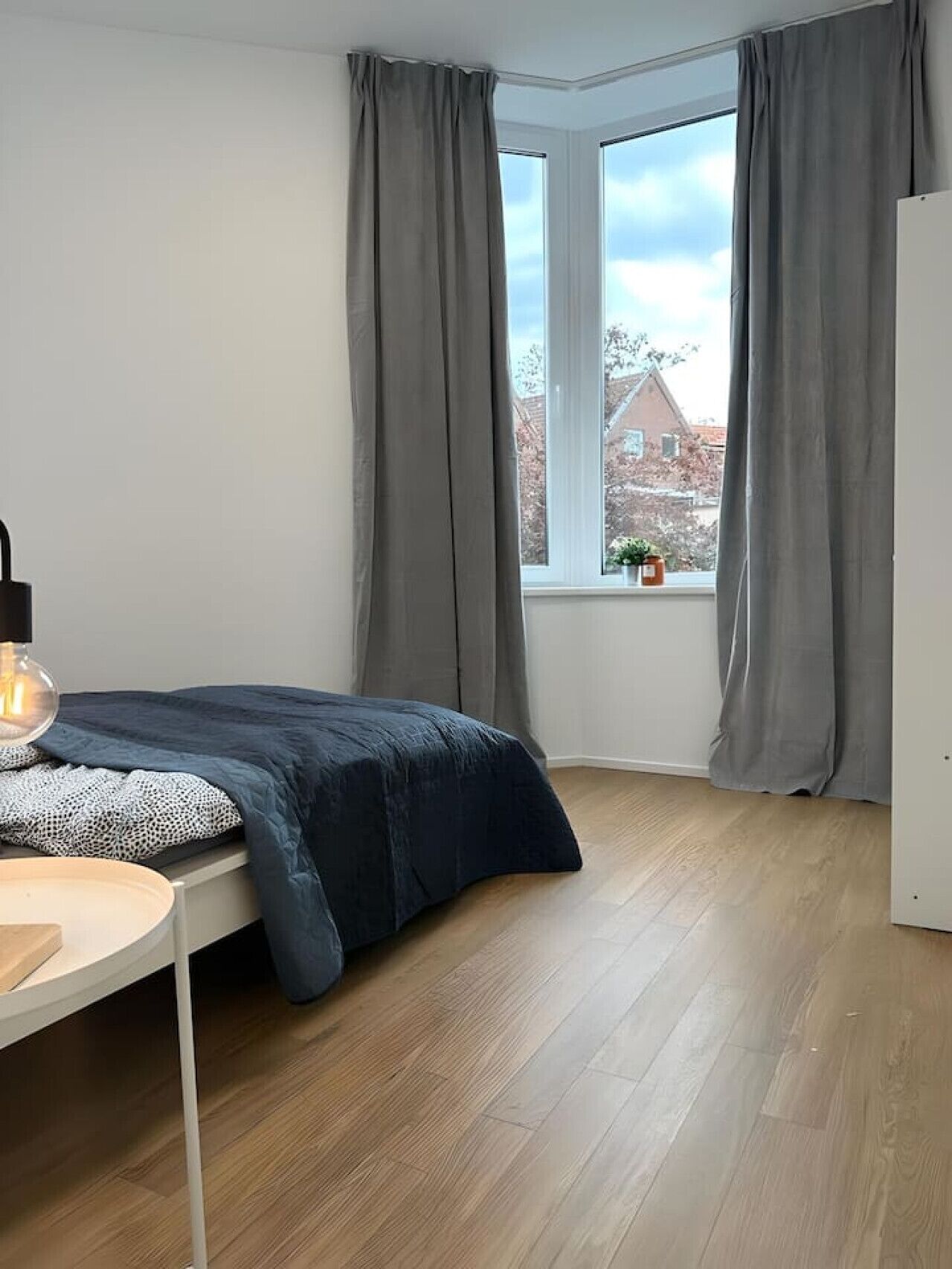 A bedroom with a bed, a bedside table, a lamp, a window with curtains, and a view of buildings outside.