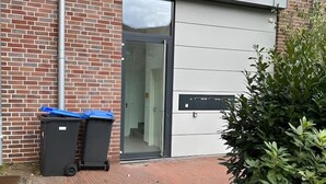 Ein Gebäude mit Backsteinfassade, einem Glastür-Eingang und zwei schwarzen Müllbehältern mit blauen Deckeln.