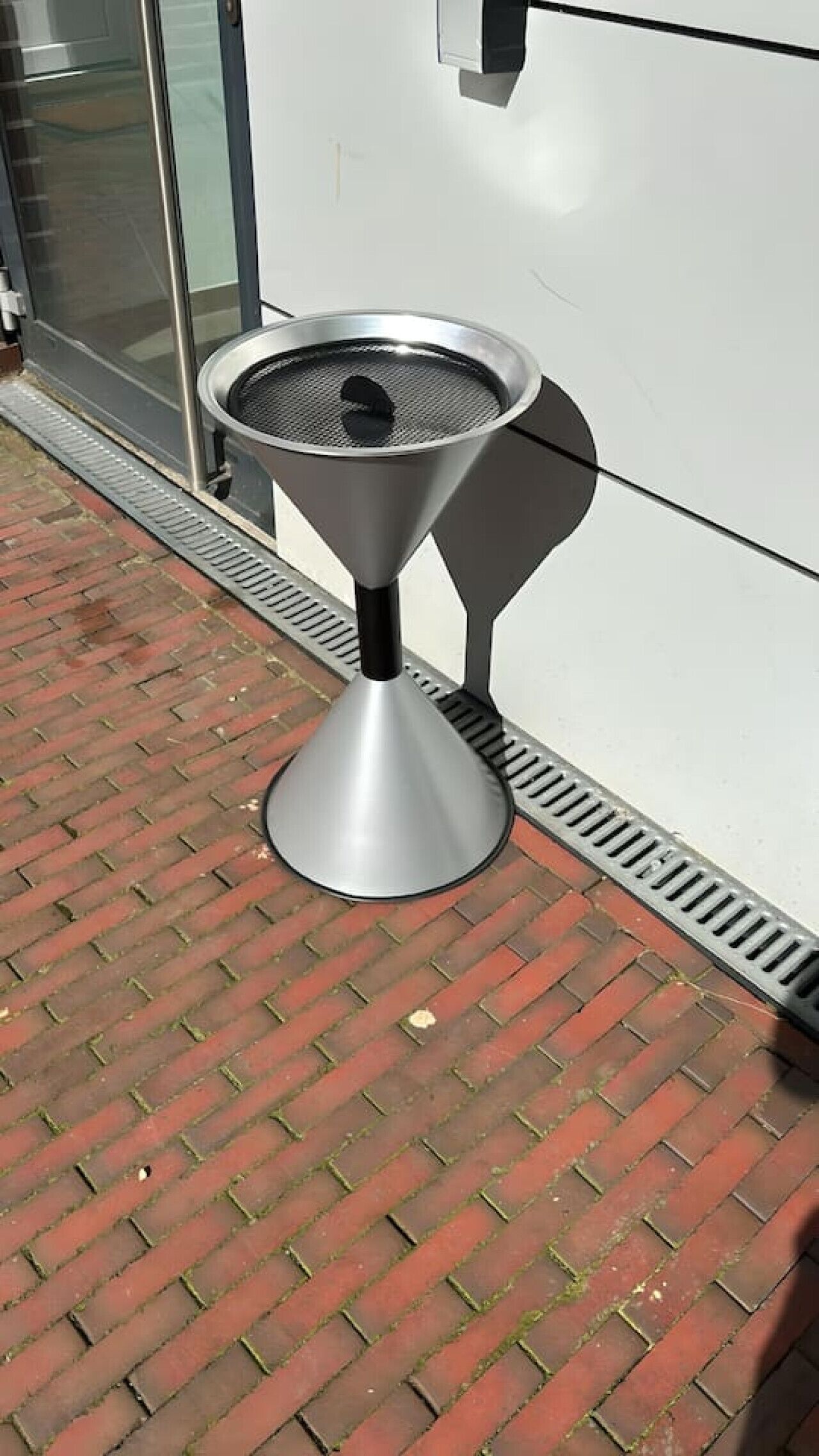 En udendørs grill i metal med en kegleformet base, placeret på en brobelagt sti ved siden af en bygning med en glasdør.