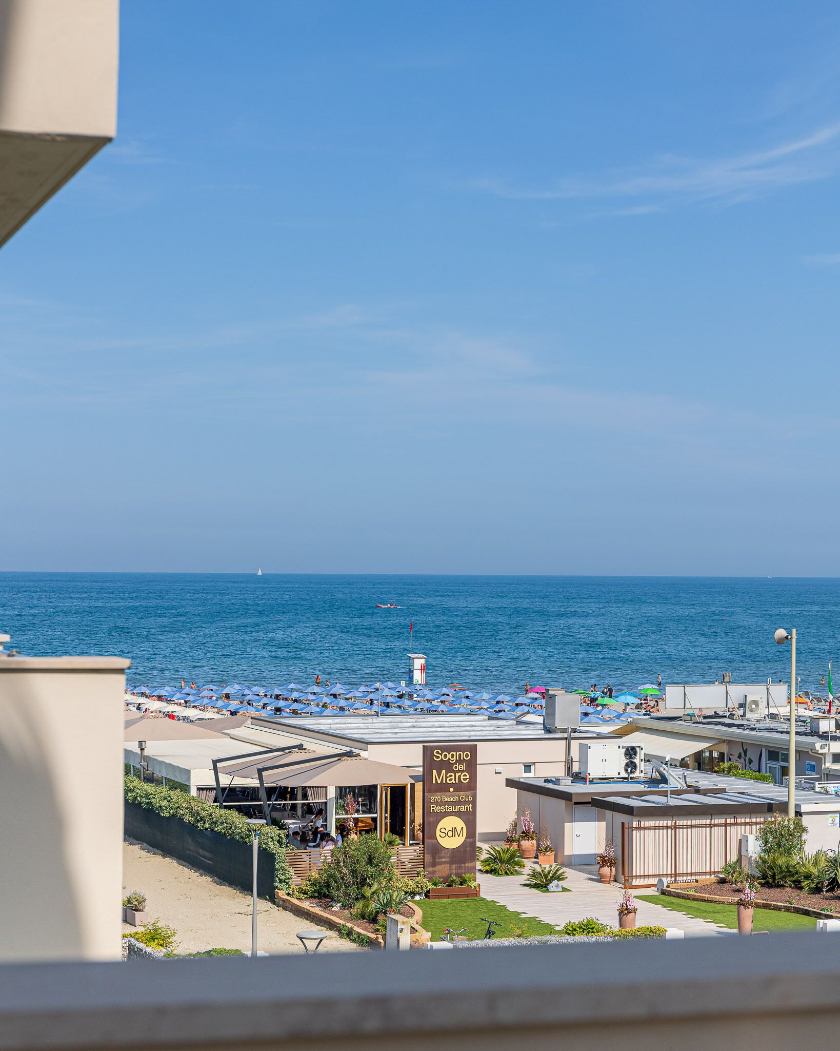 Blick auf einen Strand mit einem Restaurant namens "Sogno del Mare" und wolkenlosem, blauem Himmel.