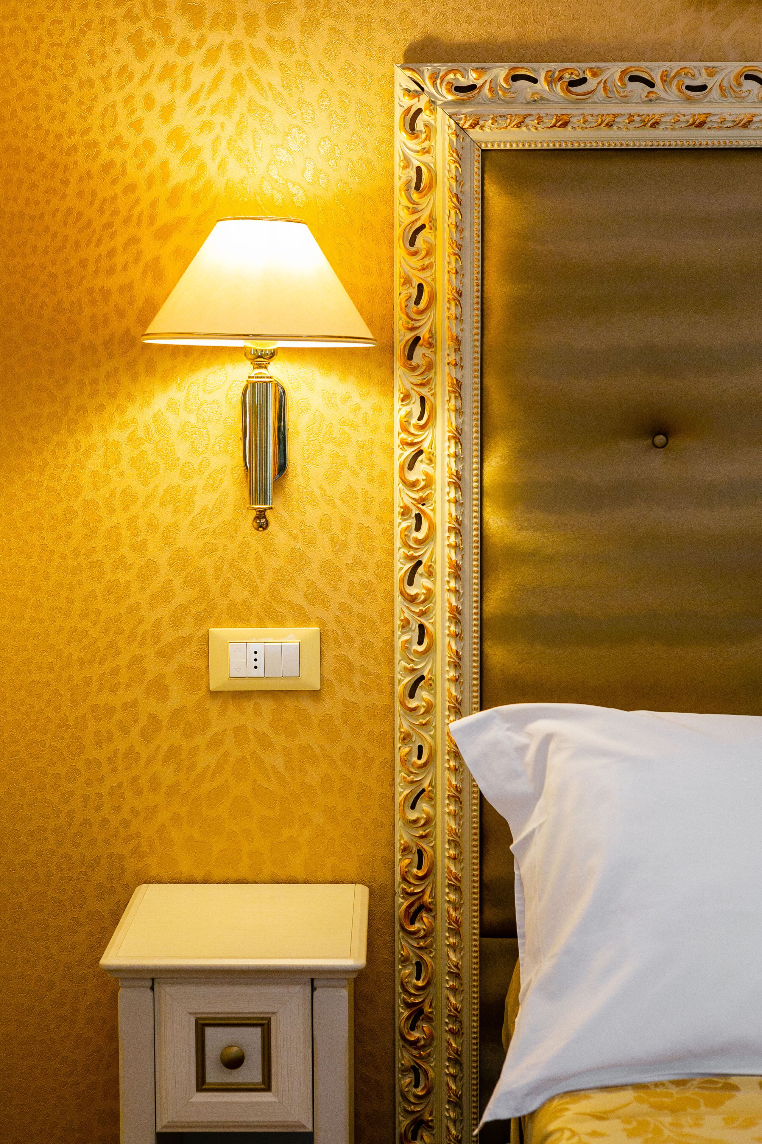 A hotel room with a gold-patterned wall, a gold-framed headboard, a bedside lamp, a nightstand, and a white pillow.