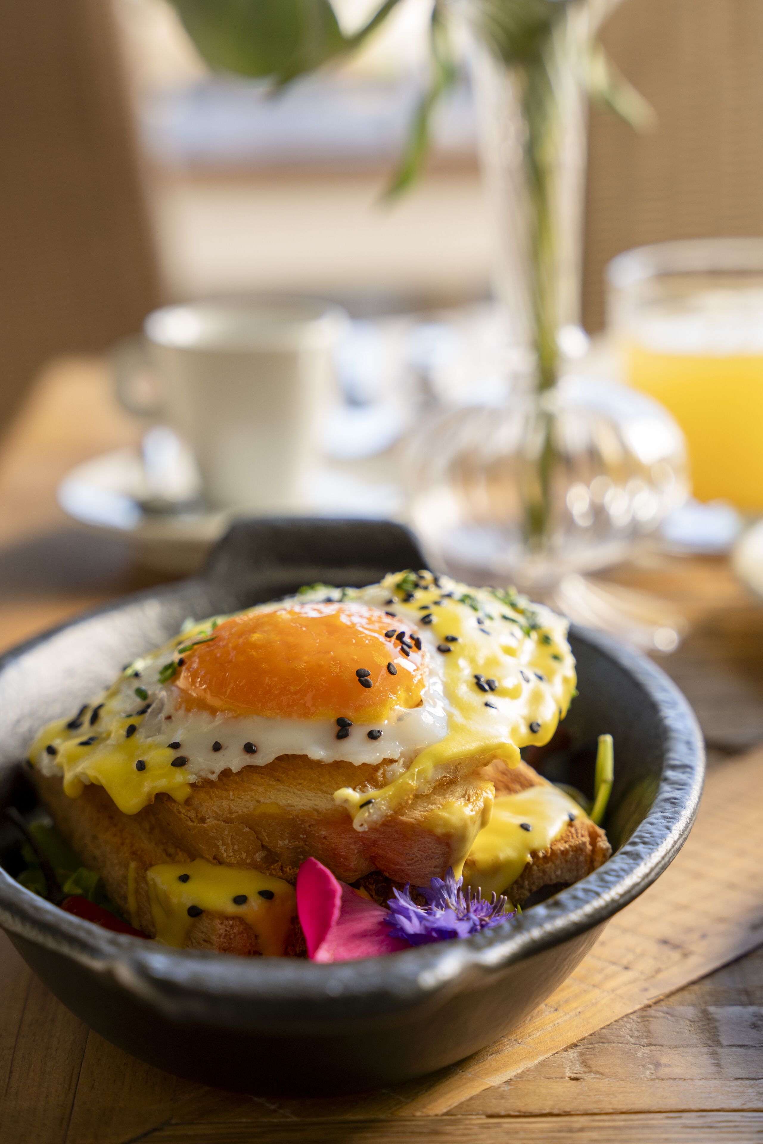 A breakfast dish with poached eggs, hollandaise sauce, and seeds on toast, served in a bowl on a wooden table.