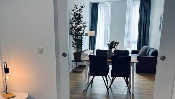 A modern, well-lit room with a wooden floor, a bed, a dining area with a table and chairs, and a living area with a sofa. - Absolute Bestlage Wohnung Artic I (Nordhorn)