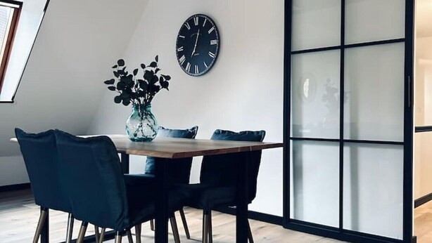A dining area with a wooden table, two chairs, a vase with flowers, and a wall clock.