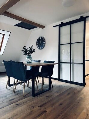 Een eetgedeelte met een houten tafel, twee stoelen, een vaas met bloemen en een wandklok.