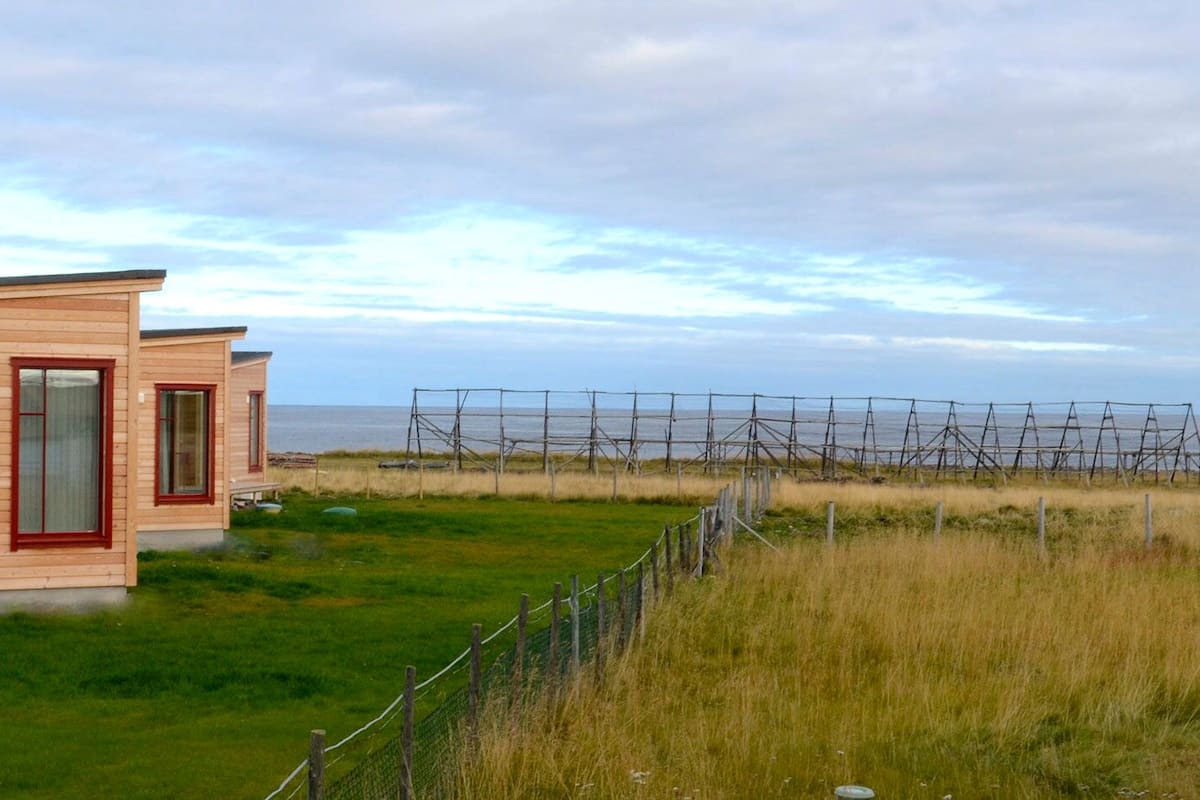 A wooden cabin with a red door and windows, situated on a grassy area with a view of the sea and power lines in the background.