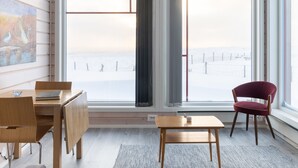 A room with a wooden desk, a chair, a small table, and a large window with a view of a snowy landscape. - Ekker Island Arctic Lodge (Vadsø)