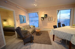 A cozy room with a sofa, armchair, and dining table, large windows with a view, and a painting on the wall.