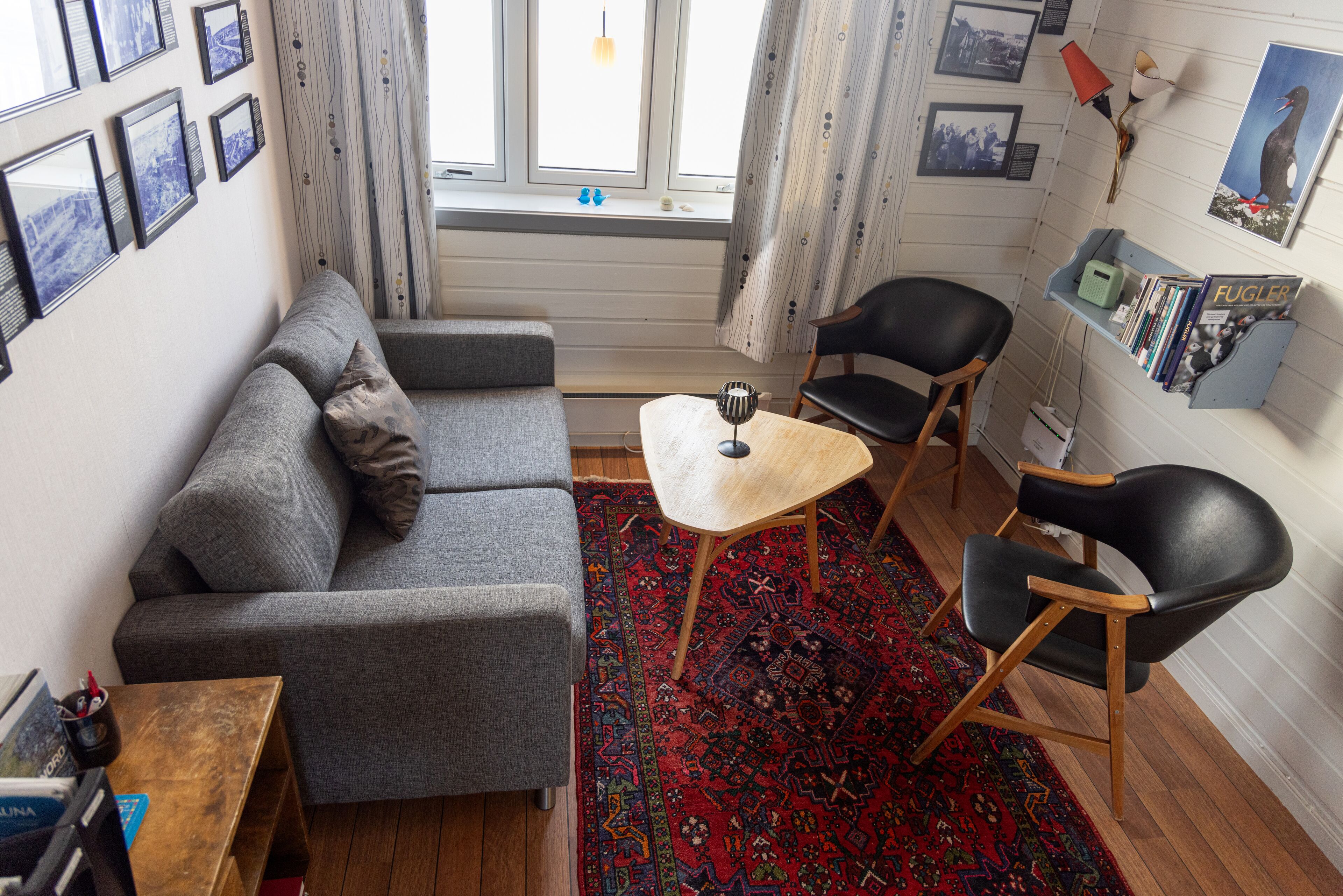 A cozy living room with a grey sofa, a small wooden table, and two black chairs. There is a red patterned rug on the floor, and a bookshelf with books and a poster on the wall.