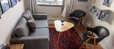 A cozy living room with a grey sofa, a small wooden table, and two black chairs. There is a red patterned rug on the floor, and a bookshelf with books and a poster on the wall.