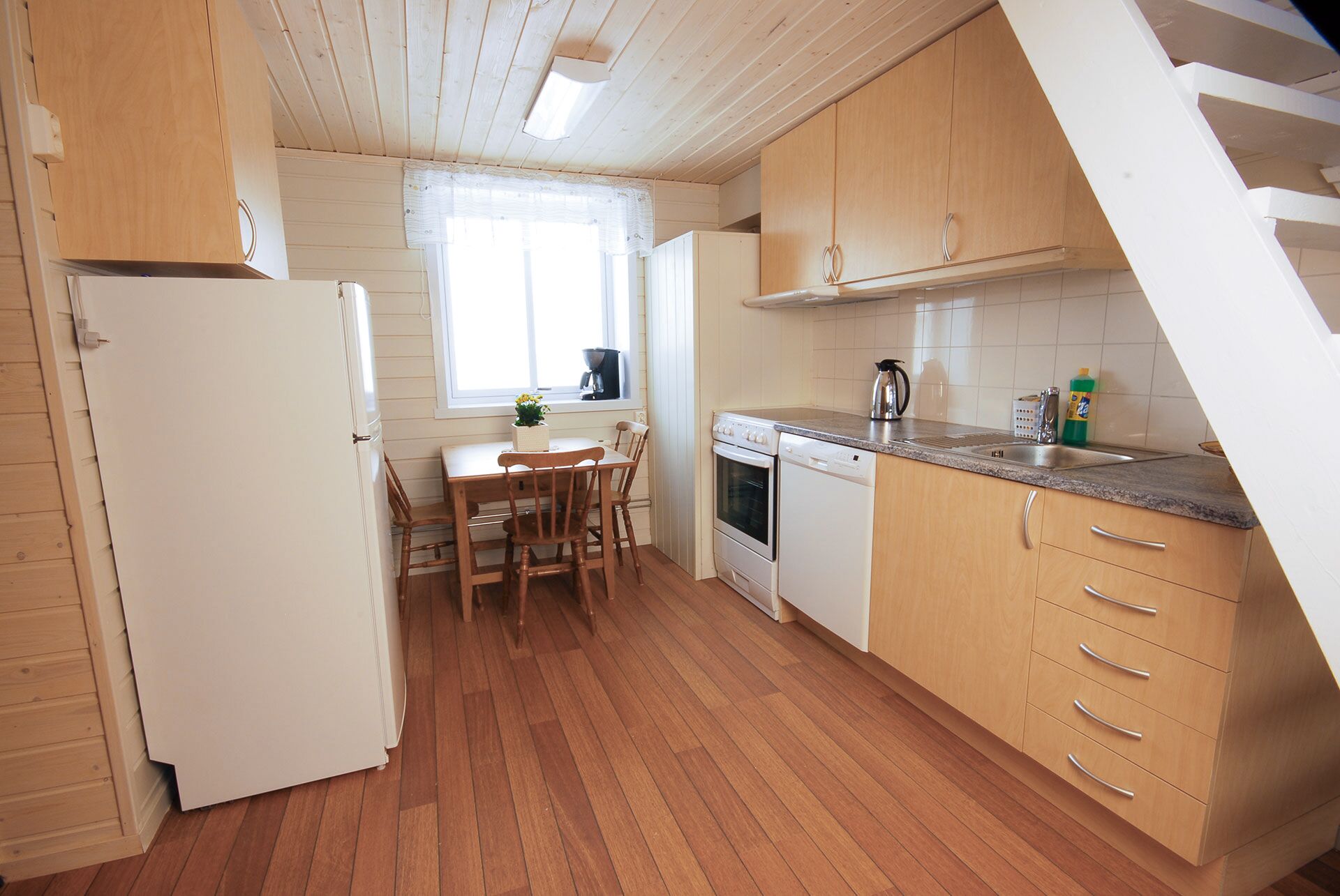 A kitchen with wooden cabinets, a white refrigerator, a dining table with chairs, a window with curtains, and a staircase.