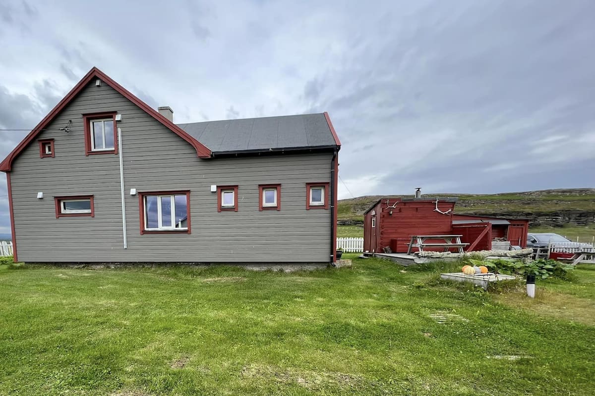 A two-story house with a grey exterior and red trim, a white picket fence, and a red shed in the background.