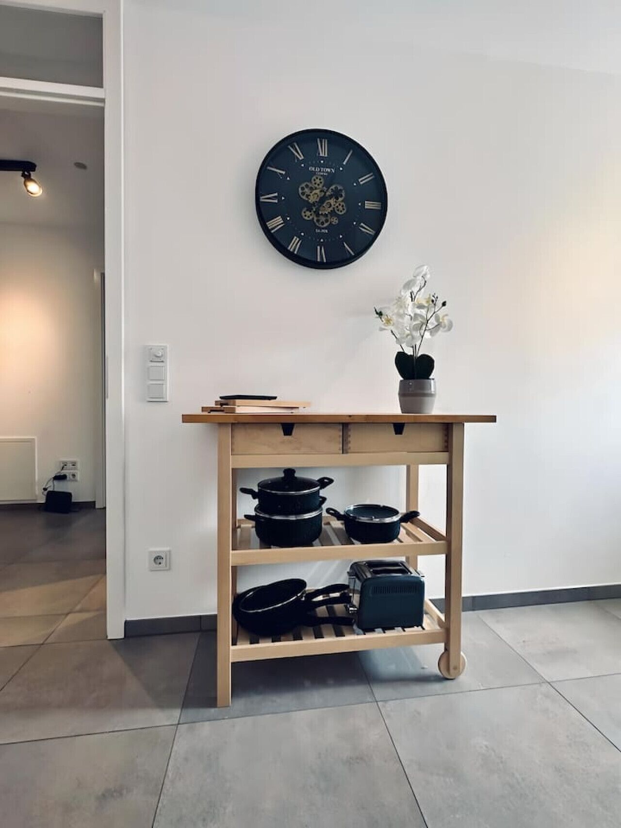 A kitchen area with a wooden shelf holding pots and pans, a clock on the wall, and a potted orchid on the shelf.