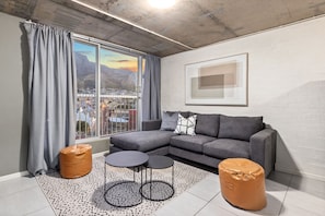 A modern living room with a grey sectional sofa, two round tables, and two ottomans. A large window offers a view of a cityscape and mountains. - The Hub 602 (Cape Town)