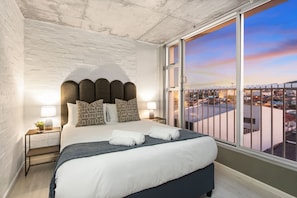 A modern hotel room with a large bed, a view of the city, and a brick wall.