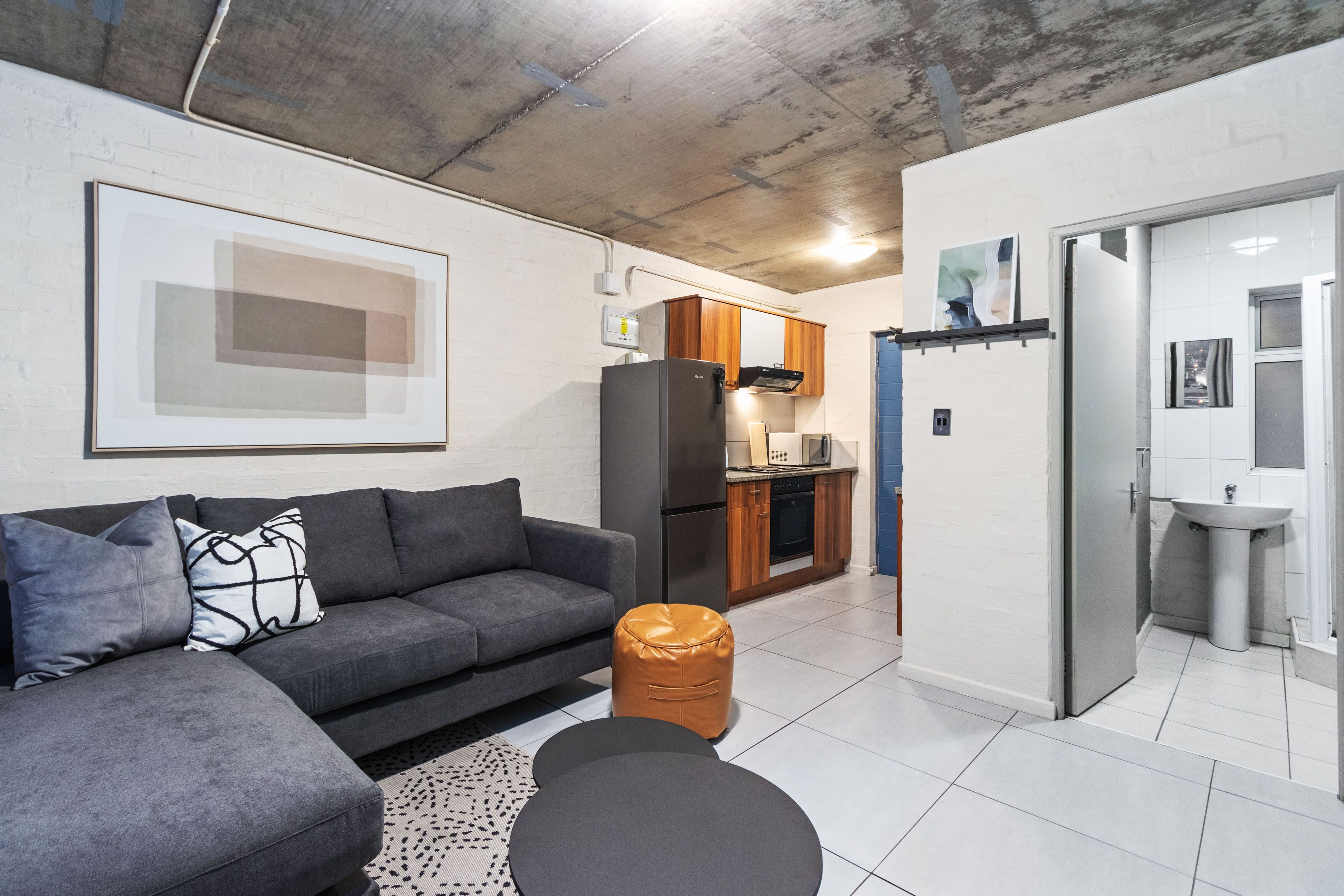 A modern apartment with a grey sectional sofa, a small round ottoman, a kitchen with wooden cabinets, and a white bathroom with a sink and mirror.
