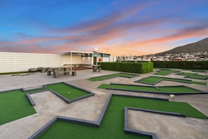 A miniature golf course with green artificial turf and concrete pathways, set against a backdrop of a cityscape and a mountain during sunset. - The Hub 602 (Cape Town)