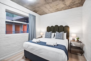 A modern hotel room with a large bed, two bedside tables with lamps, a view of an outdoor balcony, and a white brick wall. - The Hub 602 (Cape Town)