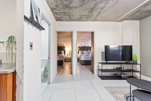 A modern living room with a flat-screen TV, a coffee table, and a sofa. There is a hallway leading to a bedroom with a bed and a nightstand. - The Hub 602 (Cape Town)
