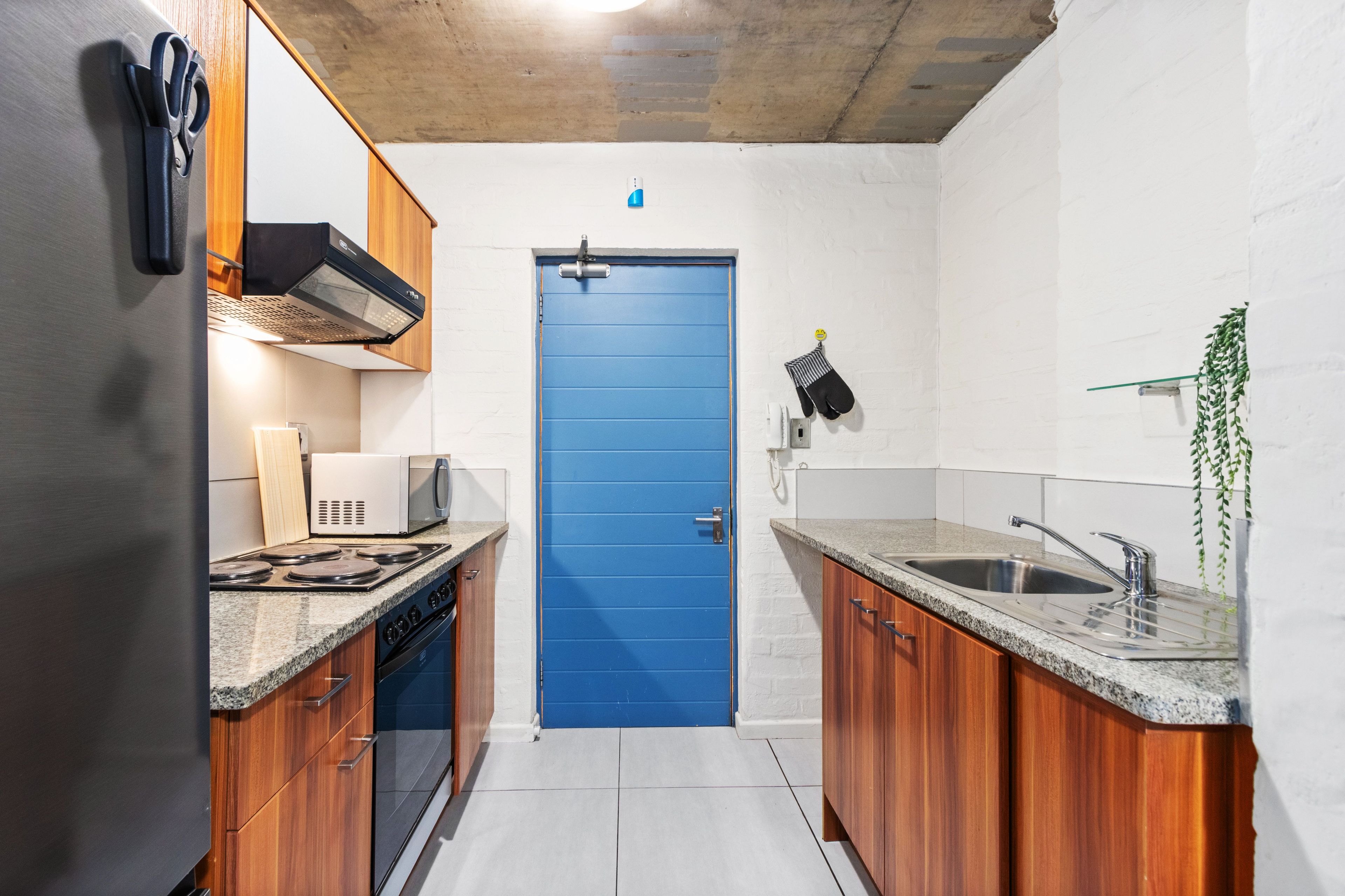 Une cuisine de taille réduite, avec des meubles de rangement en bois, une porte bleue, un four à micro-ondes et un évier.