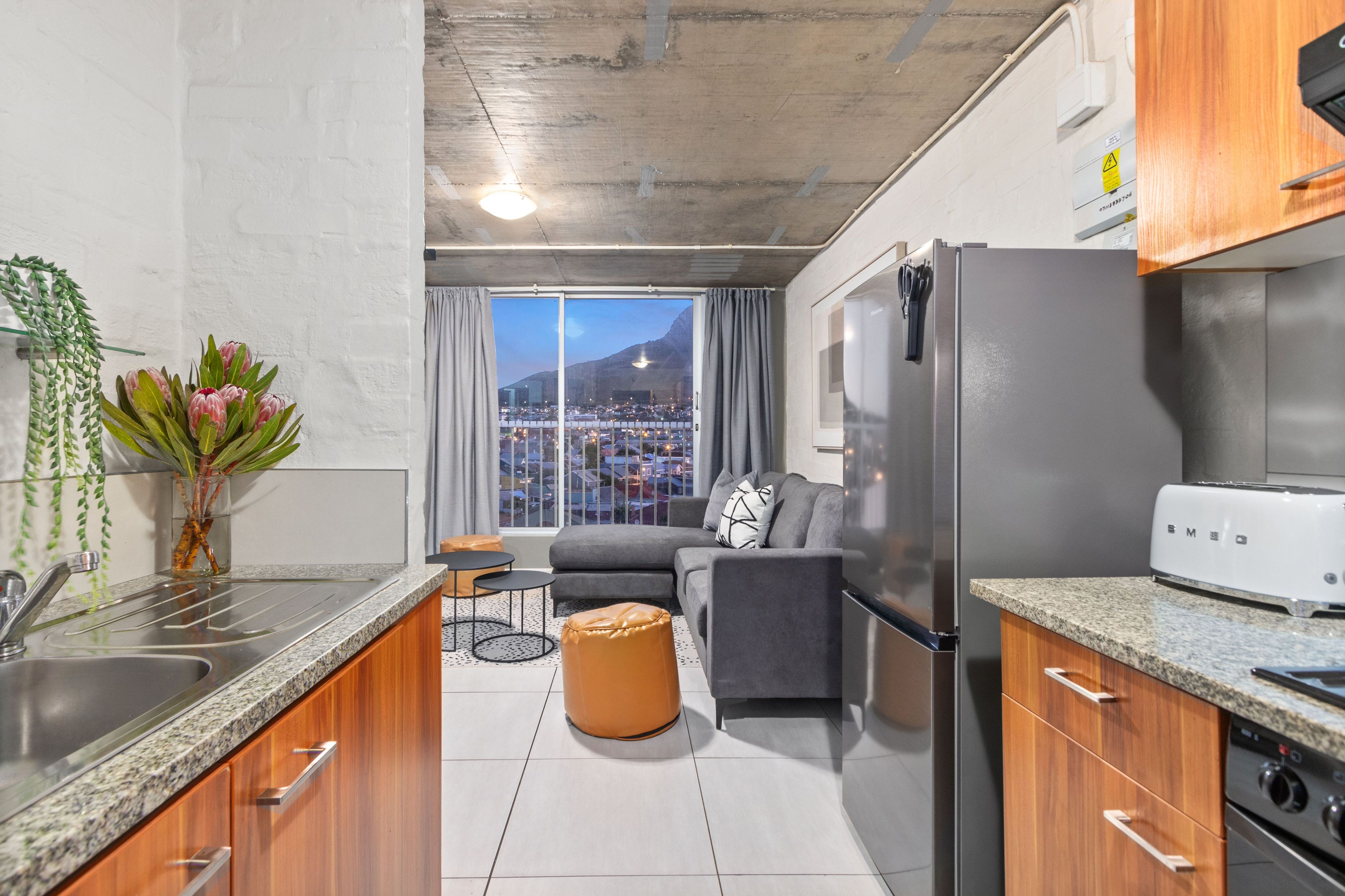 Una cocina moderna con encimeras de granito, un refrigerador de acero inoxidable y un horno empotrado. Cuenta con una zona de comedor con un sofá gris y una pequeña mesa redonda. Un ventanal ofrece vistas a un paisaje urbano y una montaña.