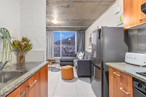 A modern kitchen with granite countertops, a stainless steel refrigerator, and a built-in oven. There is a dining area with a gray sofa and a small round table. A large window offers a view of a cityscape and a mountain. - The Hub 602 (Cape Town)