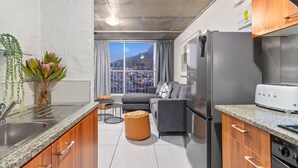 A modern kitchen with granite countertops, a stainless steel refrigerator, and a built-in oven. There is a dining area with a gray sofa and a small round table. A large window offers a view of a cityscape and a mountain. - The Hub 602 (Cape Town)