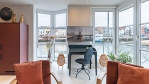 Ein modernes Wohnzimmer mit einem Klavier, einem großen Fenster mit Blick auf die Stadt und einer gemütlichen Sitzecke.